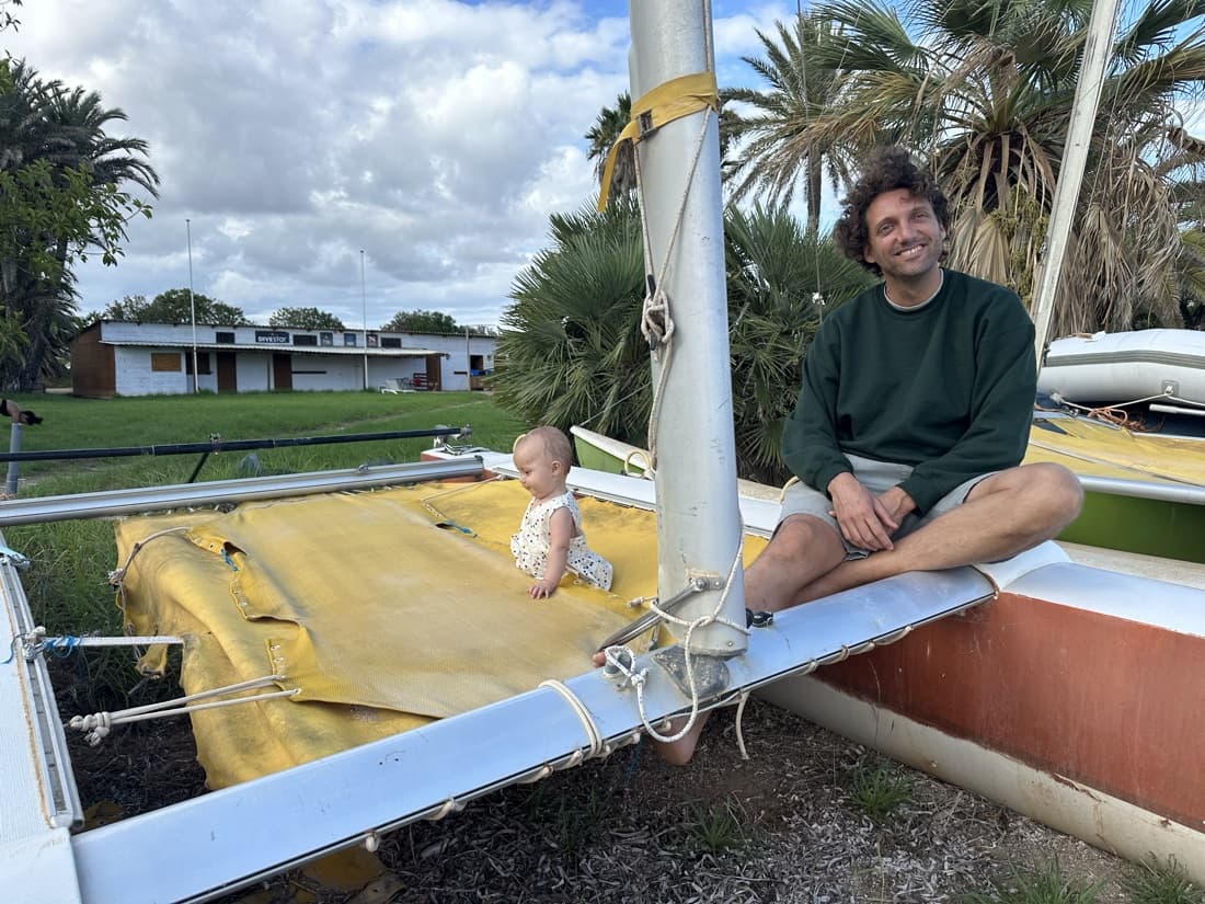 A father sitting on a beached catamaran with his baby crawling on the trampoline