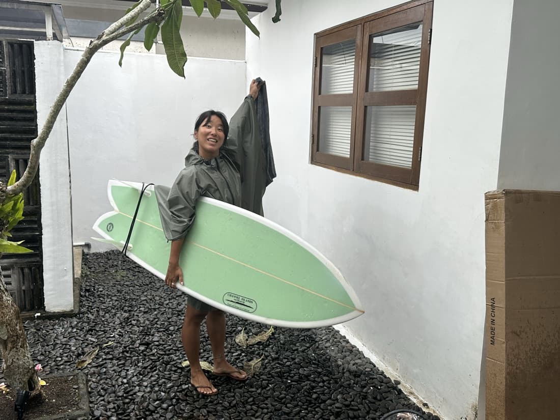 A smiling surfer in a rain poncho holding a mint-green surfboard